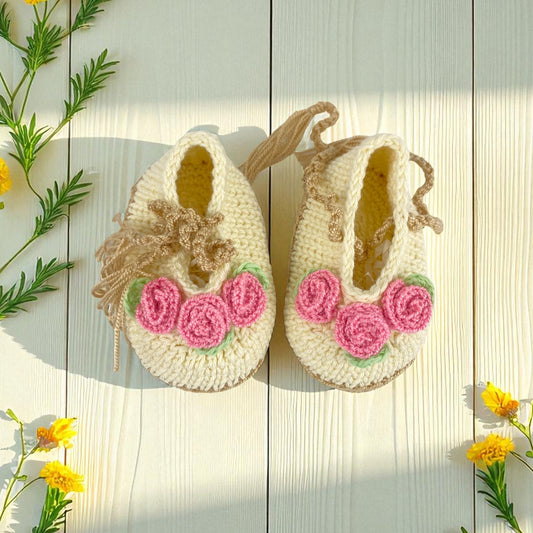 Pair of crocheted baby shoes with pink flowers on a wooden surface with flowers around.