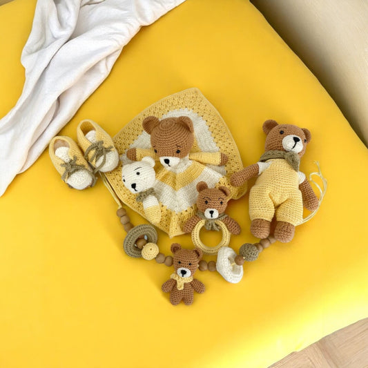Collection of teddy bear-themed baby items including a blanket, shoes, and rattle on a yellow surface.