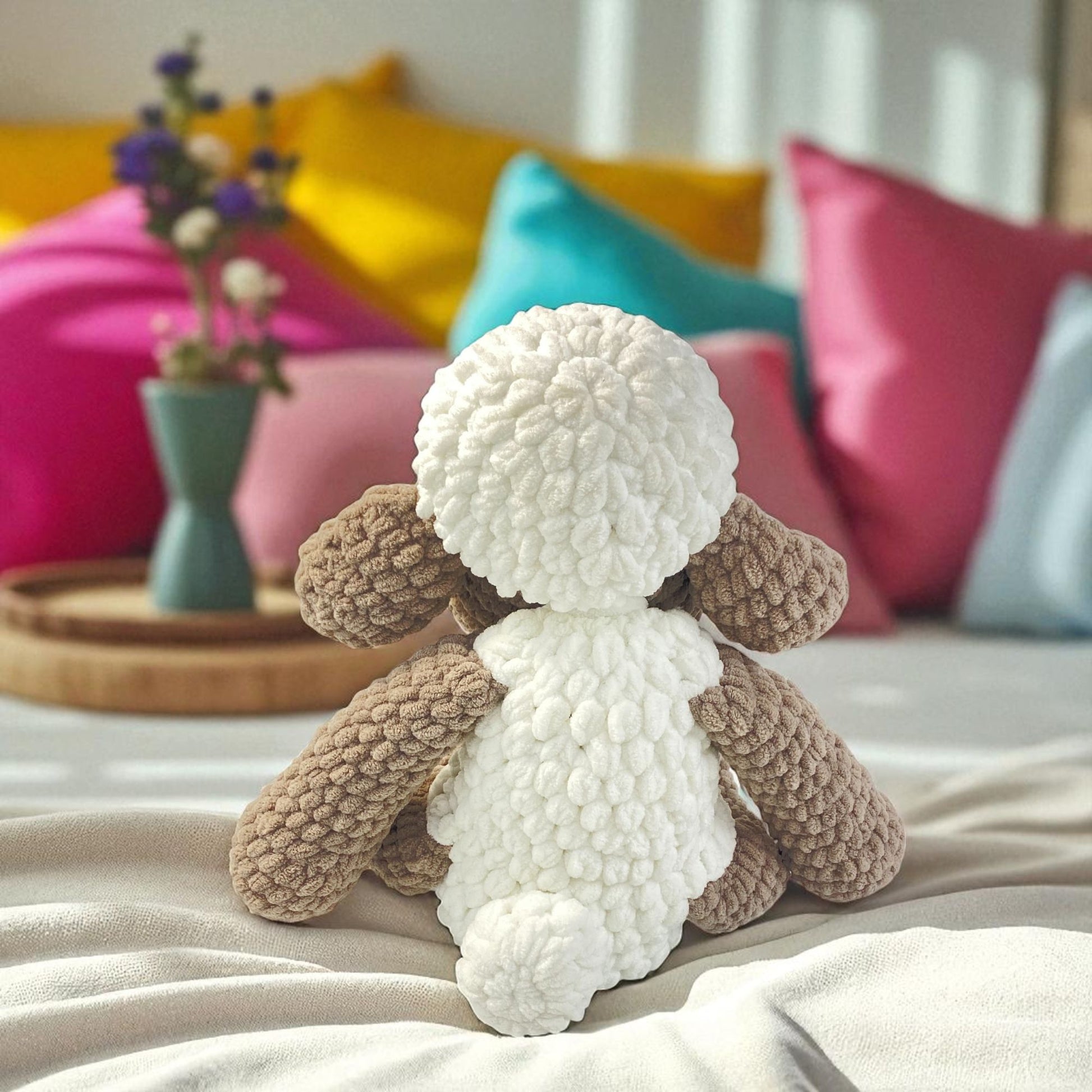 Knitted sheep toy on a bed with colorful pillows and a vase in the background