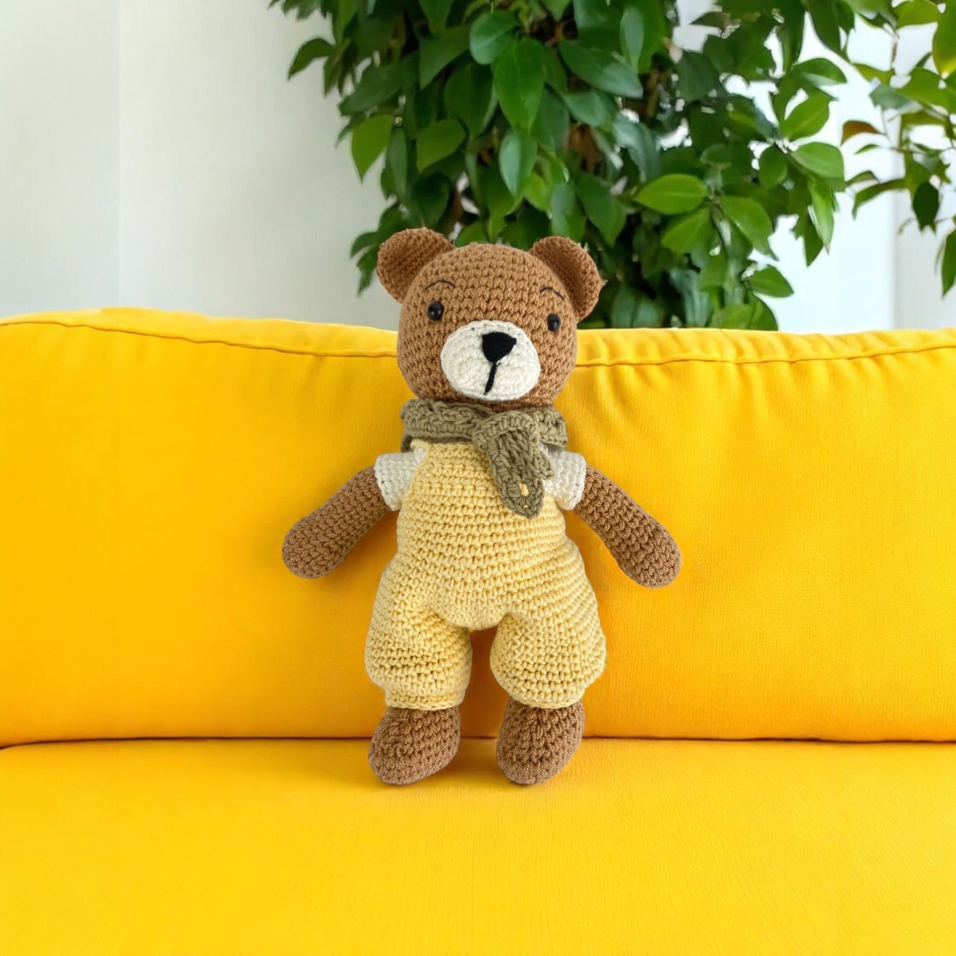 Crocheted teddy bear with a scarf on a yellow couch with a plant in the background