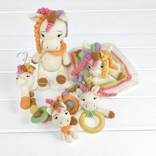 Collection of crochet  crib toys including a unicorn, a rattle, and a blanket on a white wooden background.